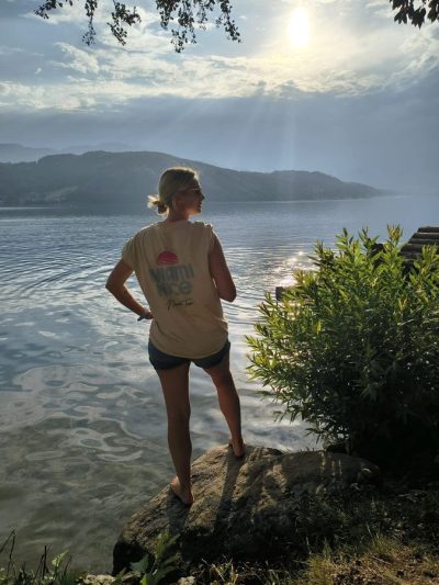 Frau im Miami-Nice-Shirt steht im Gegenlicht am Seeufer